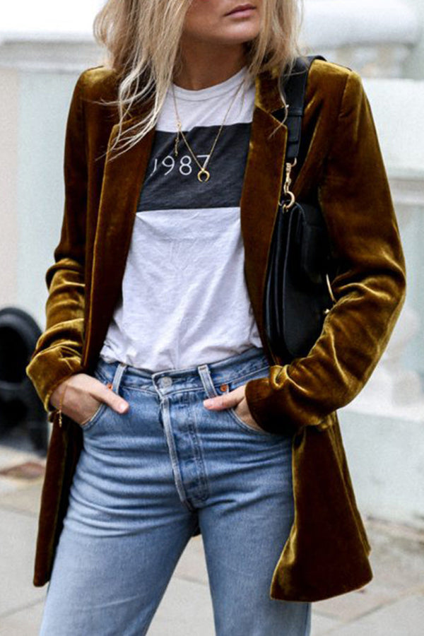 Une femme porte un blazer en velours moutarde, une chemise blanche avec un imprimé noir, et un jean bleu clair. Elle tient un sac à main noir et affiche une tenue élégante et décontractée.