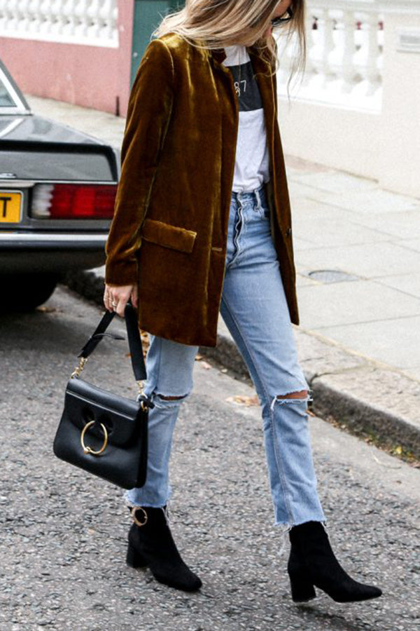 Une femme marche dans la rue en portant un blazer en velours jaune moutarde, un sac à main noir et des bottines noires. Elle porte un t-shirt blanc et un jean bleu déchiré, affichant un look moderne et décontracté.