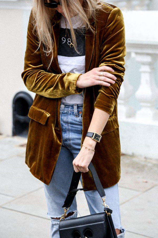 Une femme porte un blazer en velours de couleur moutarde, un t-shirt blanc avec un imprimé noir, et un jean bleu déchiré. Elle tient un sac à main noir et porte des bijoux discrets. Elle est capturée en pleine rue, avec une allure décontractée et élégante.