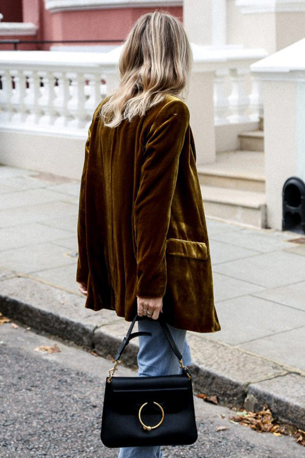 Une femme vue de dos porte un blazer en velours jaune moutarde, avec un sac à main noir à l'épaule. Elle marche sur le trottoir, portant un jean bleu délavé, affichant un look élégant et décontracté.