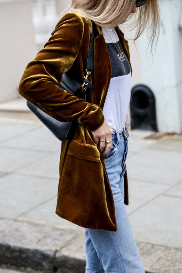 Une femme porte un blazer en velours jaune moutarde, une chemise blanche avec un imprimé noir et un jean bleu. Elle tient un sac à main noir et affiche un look chic et décontracté en marchant dans la rue.