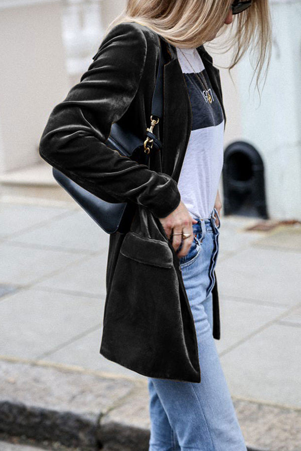 Une femme porte un blazer en velours noir, une chemise blanche avec un imprimé noir et un jean bleu. Elle tient un sac à main noir et affiche un look élégant et décontracté en marchant dans la rue.
