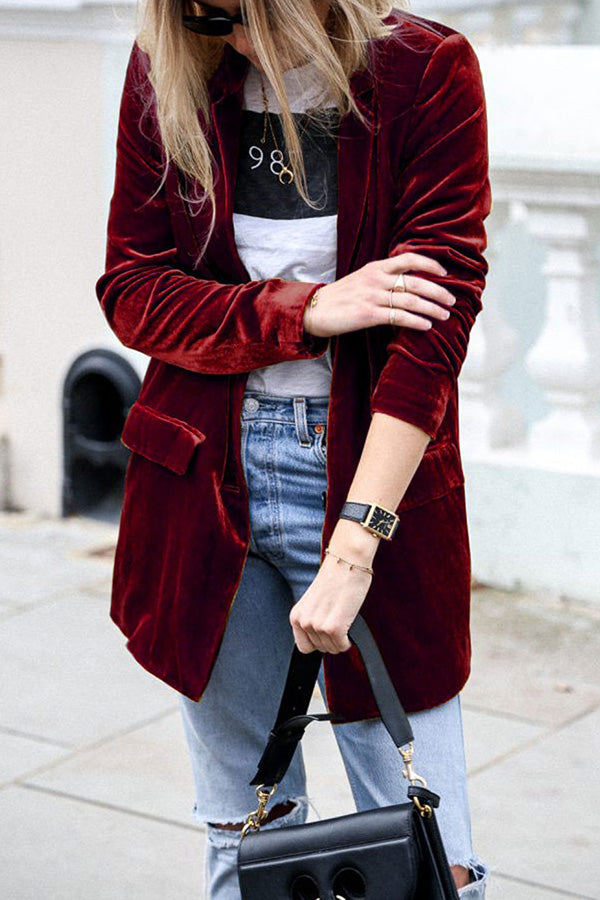 Une femme portant un blazer en velours rouge avec des manches retroussées, une chemise blanche avec des rayures noires, et un jean bleu clair. Elle tient un petit sac à main noir, affichant un look élégant et décontracté.
