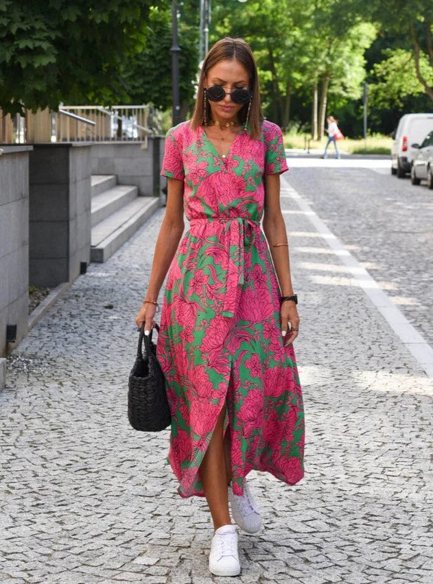 Robe midi Layla pour femme au style moderne, col en V et imprimé floral rose et vert, coupe fluide avec ceinture à la taille, portée avec des baskets blanches et un sac noir pour un look élégant et décontracté en extérieur.