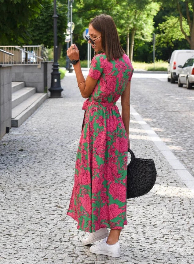 Robe midi Layla pour femme, vue de dos, col en V et coupe moderne, imprimé floral rose sur fond vert, taille ajustée avec ceinture, tissu fluide, associée à des baskets blanches et un sac noir pour un look élégant et décontracté.