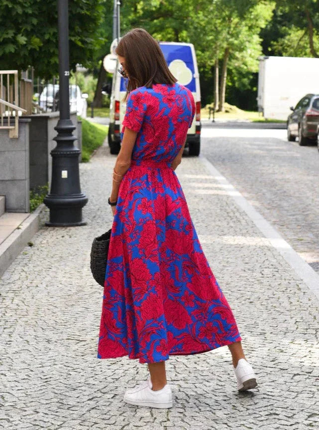 Robe midi Layla pour femme, vue de dos, col en V et coupe moderne, imprimé floral rouge sur fond bleu, taille ceinturée et jupe fluide, portée avec des baskets blanches et un sac noir pour un look chic et décontracté.