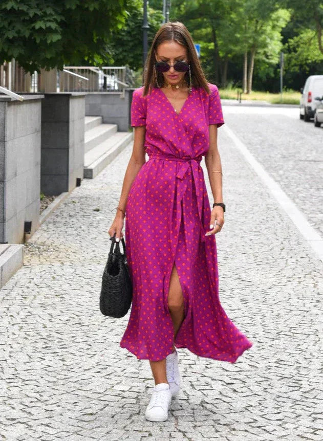 Robe midi Layla pour femme, col en V et coupe moderne, coloris violet avec motifs à pois, taille ceinturée et tissu fluide, portée avec des baskets blanches et un sac noir pour un look chic et décontracté en extérieur.