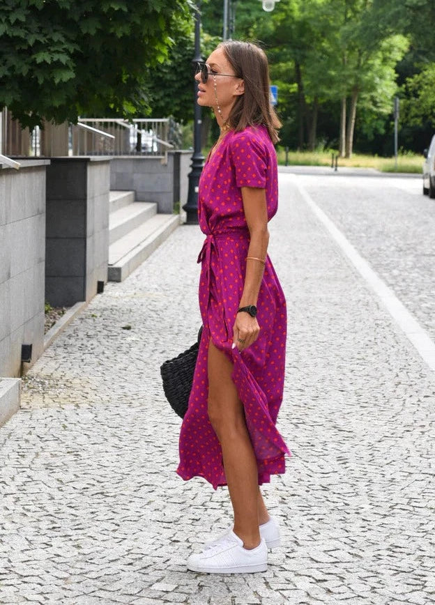 Robe midi Layla pour femme, vue de profil, col en V et coupe moderne, coloris violet à pois avec fente élégante, taille ceinturée et tissu fluide, portée avec des baskets blanches et un sac noir pour un style chic et décontracté.