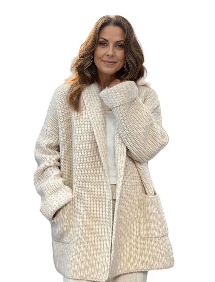Femme portant un long cardigan en maille beige, au style confortable et élégant, posant dans un intérieur lumineux avec des vêtements suspendus en arrière-plan.