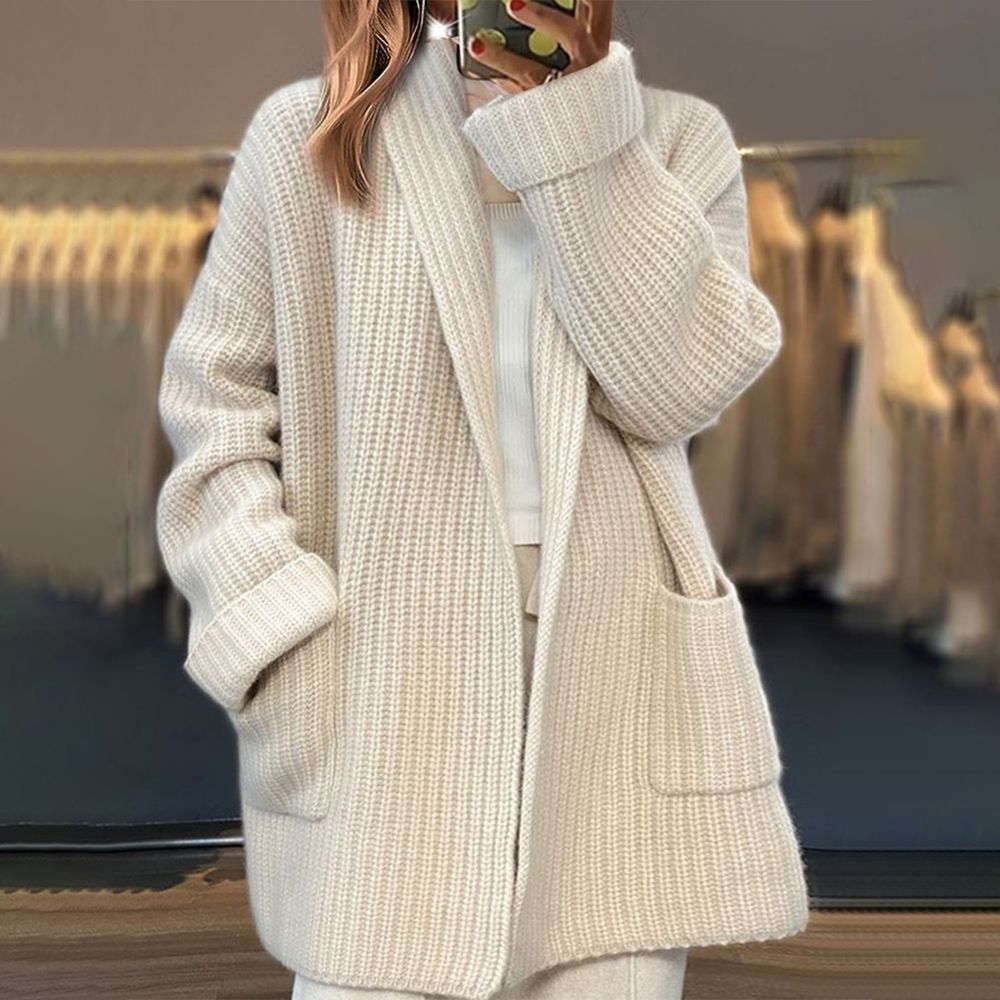 Femme prenant une photo en portant un long cardigan en maille beige, avec une coupe ample et des poches avant, dans un intérieur où des vêtements sont suspendus en arrière-plan.