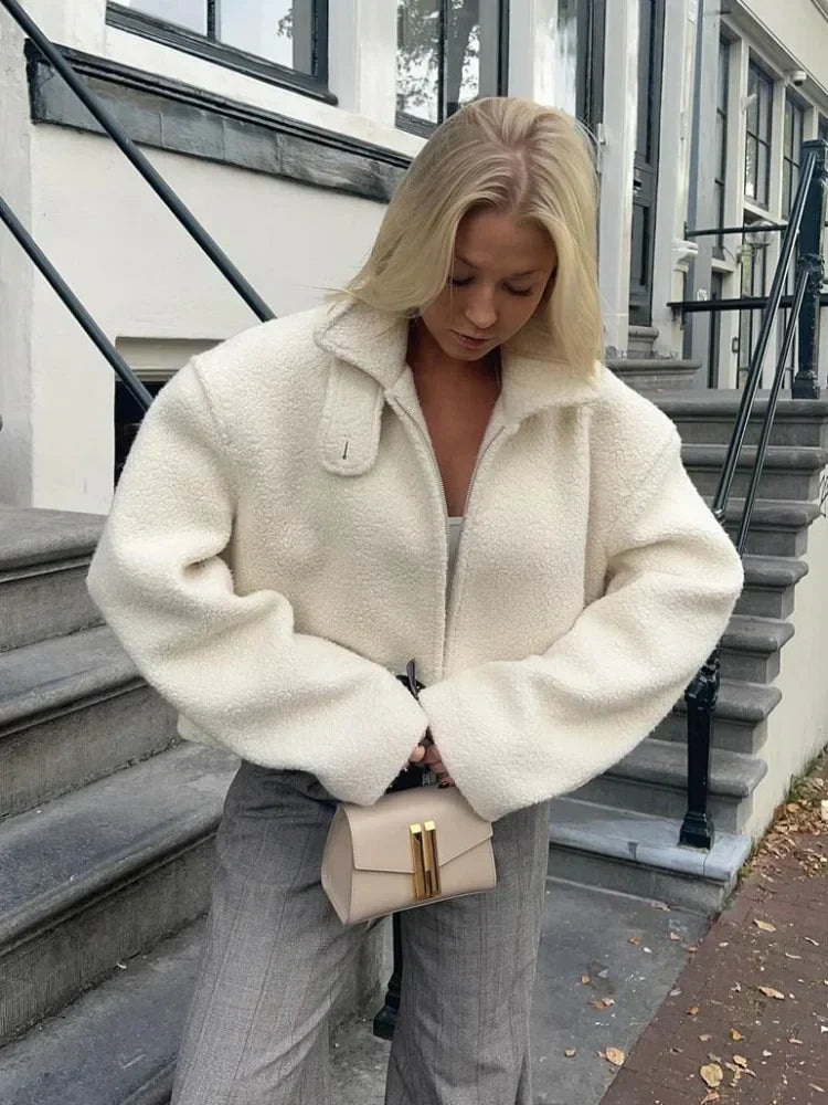 Une femme porte une veste polaire blanche avec une coupe ample et moderne, parfaitement adaptée pour l’hiver. Elle la porte avec un pantalon à rayures gris et tient un petit sac beige aux détails dorés. Elle est debout sur des marches en pierre, avec une posture détendue, ses cheveux blonds légèrement ondulés tombant sur ses épaules. Son regard est dirigé vers le bas, créant une ambiance chic et décontractée.