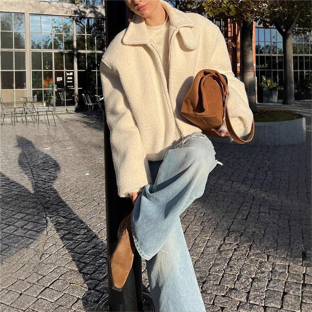 Une femme est assise dehors, portant une veste polaire blanche au style décontracté avec un col replié. Elle a un jean bleu délavé et des bottines beige, et tient un sac marron en cuir. La lumière du soleil illumine la scène, créant une ambiance chaleureuse et élégante. L'arrière-plan montre un environnement urbain avec des chaises en extérieur et des arbres, ajoutant à l'atmosphère détendue de la journée.