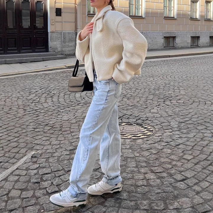 Une femme marche dans une rue pavée, portant une veste polaire blanche au style décontracté avec un col légèrement replié. Elle a un jean bleu clair et des baskets blanches. Elle tient un sac à main beige à la main. L’arrière-plan présente une architecture élégante, ajoutant une touche urbaine à son look casual et confortable, parfait pour une sortie hivernale.