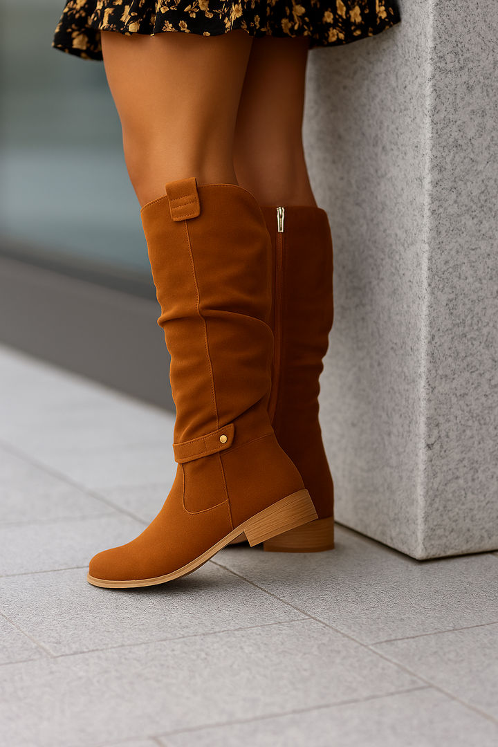 Jambes d’une femme portant des bottes montantes en daim camel, posée contre un mur en pierre. Les bottes au design moderne, dotées d’une tige souple, d’une bride décorative et d’un talon bas en bois, complètent une tenue féminine avec une jupe noire à motifs floraux.