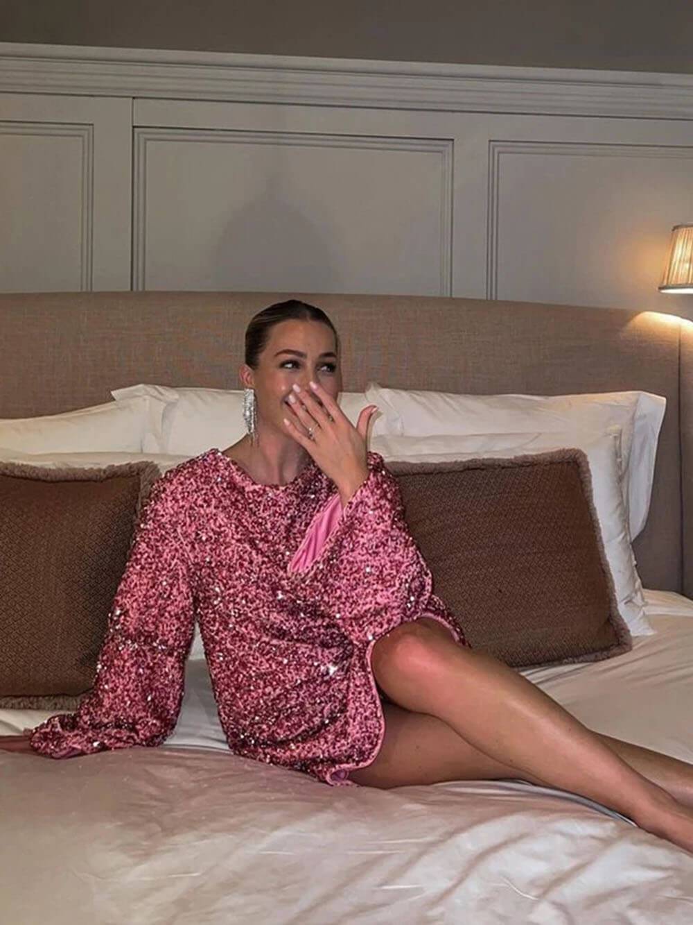 Une femme assise sur un lit, portant une robe rose à sequins avec un nœud en satin rose à l'arrière. Elle sourit et se touche légèrement le visage avec sa main gauche. Le décor montre un lit élégant avec des oreillers et des lumières douces.