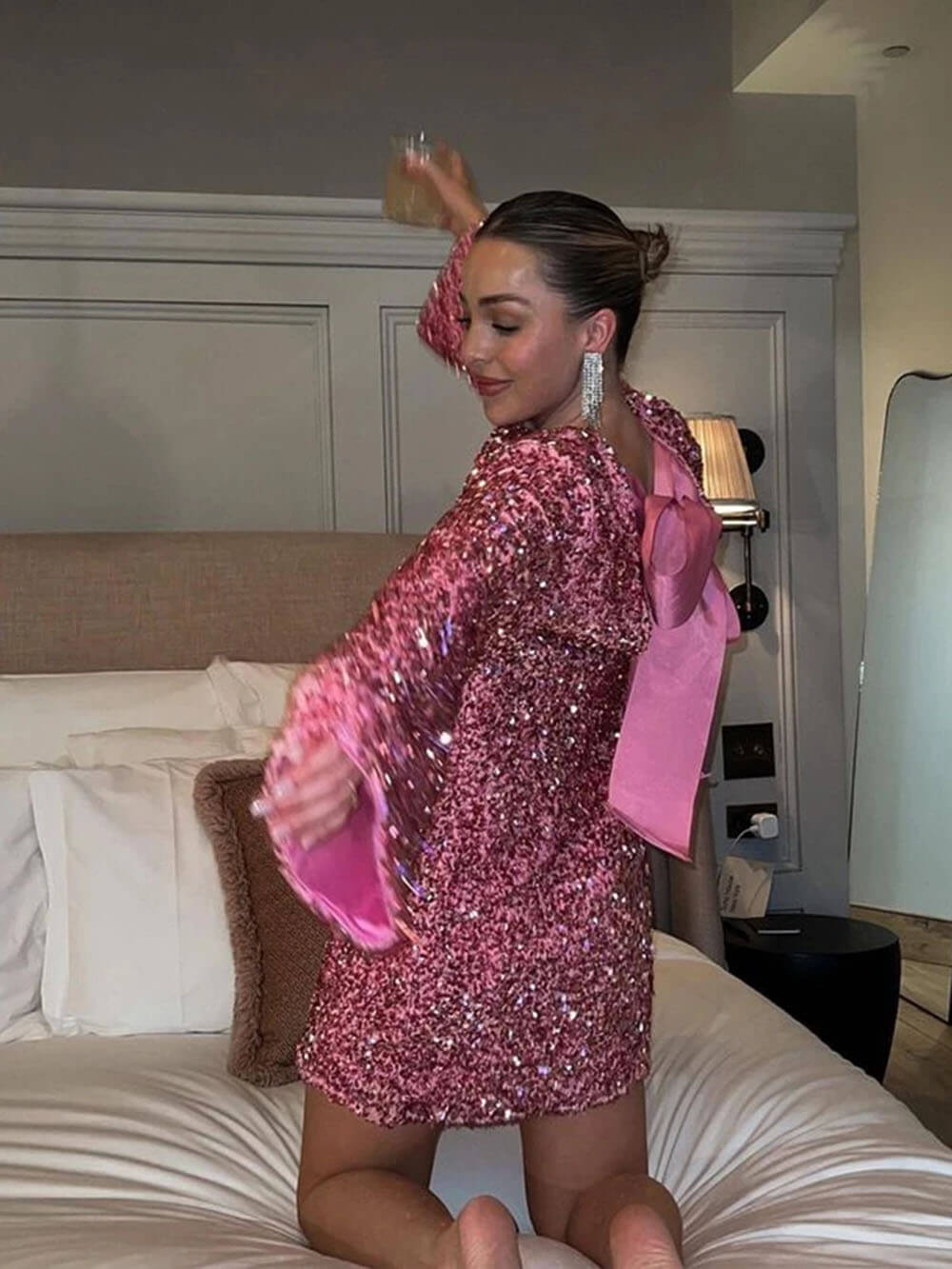 Une femme danse avec un verre à la main, portant une robe rose à sequins. Le dos de la robe est décoré d'un grand nœud en satin rose. Elle sourit et se trouve sur un lit avec des oreillers dans un cadre élégant.
