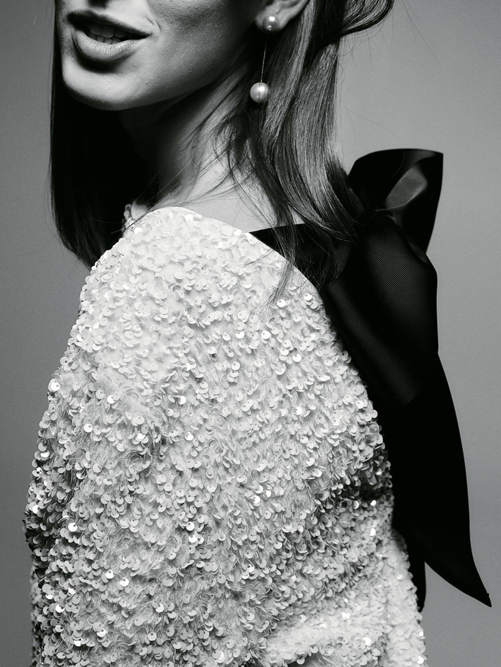 Un gros plan d'une femme souriante, portant une robe à sequins blancs avec un dos ouvert et un nœud noir. Elle porte également des boucles d'oreilles perlées, ajoutant une touche d'élégance.