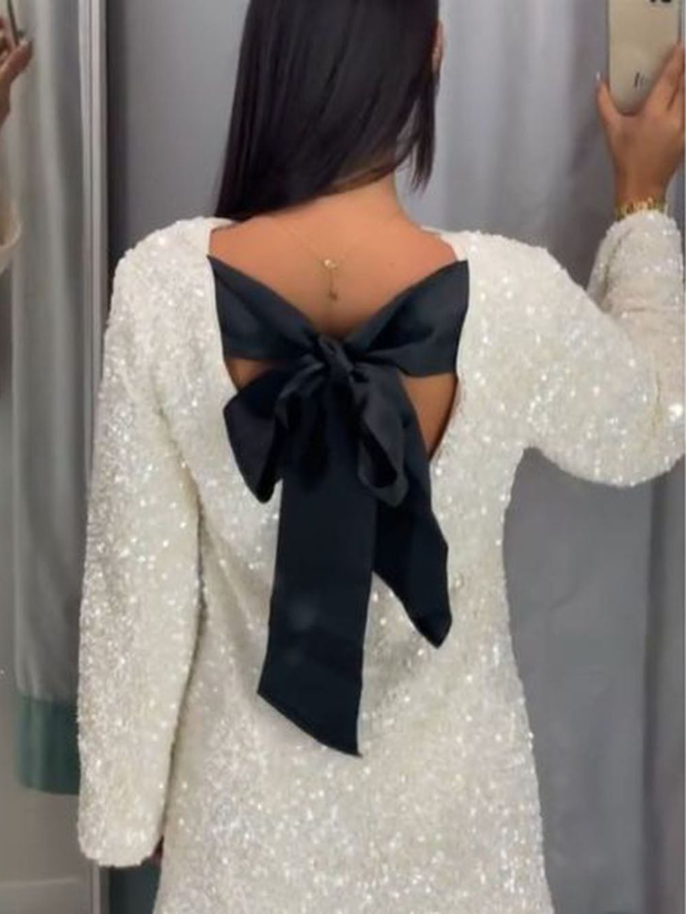 Vue arrière d'une femme portant une robe blanche à sequins avec un grand nœud noir attaché au dos. Elle est photographiée dans un miroir, mettant en valeur la silhouette élégante et le détail du nœud.