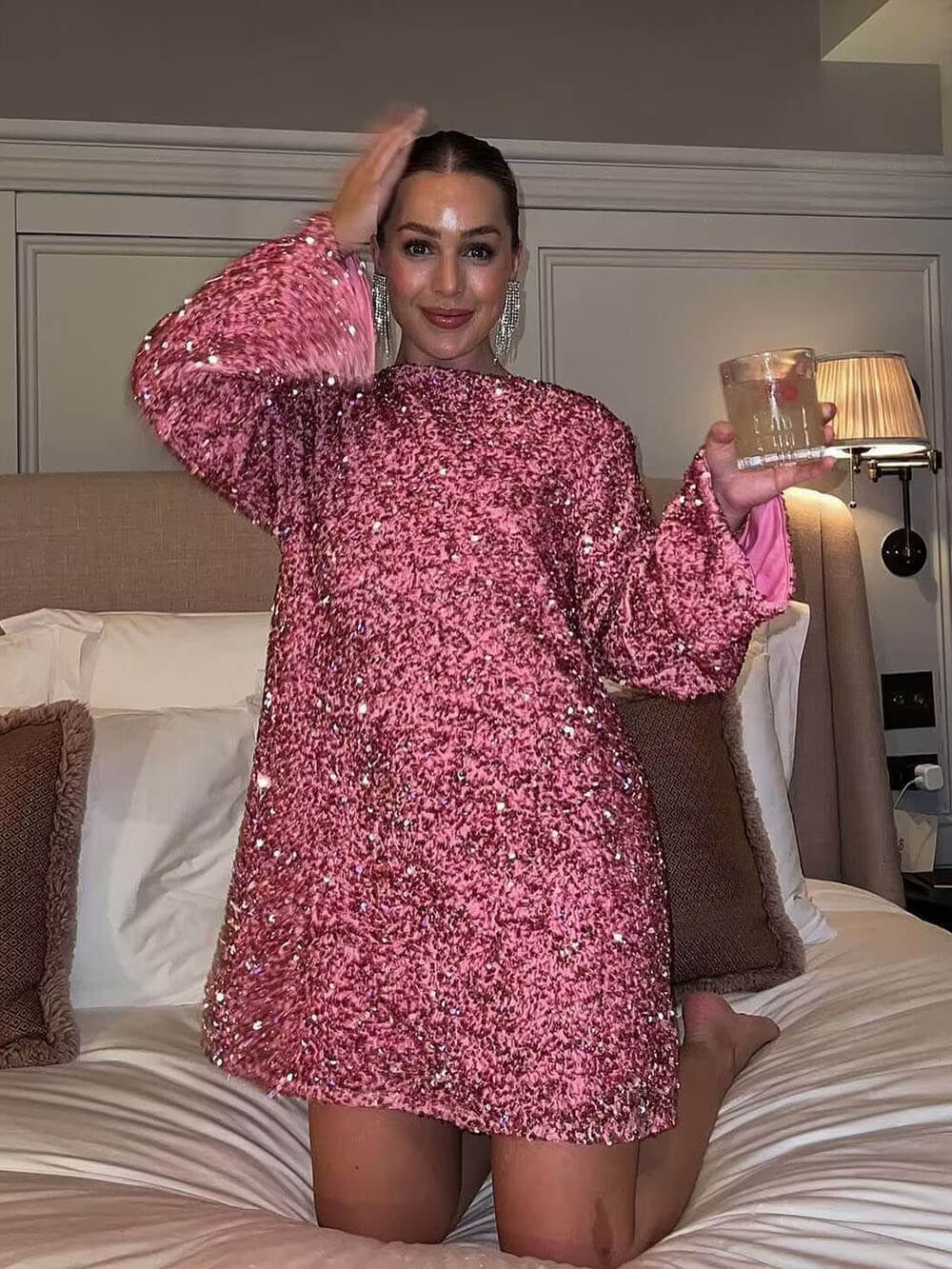 Une femme portant une robe rose à sequins, avec des manches longues évasées. Elle pose avec un verre dans une main, debout sur un lit dans une chambre. La robe brille sous l'éclairage doux, mettant en valeur la texture et la coupe élégante de la tenue.