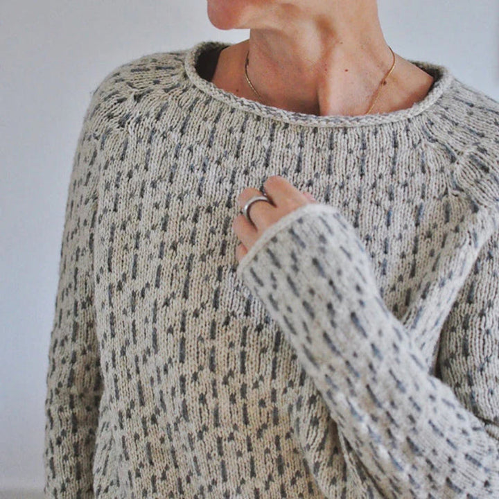 Gros plan sur une femme portant un pull en maille clair à col rond, avec un motif texturé subtil et des manches longues, dans un intérieur lumineux.