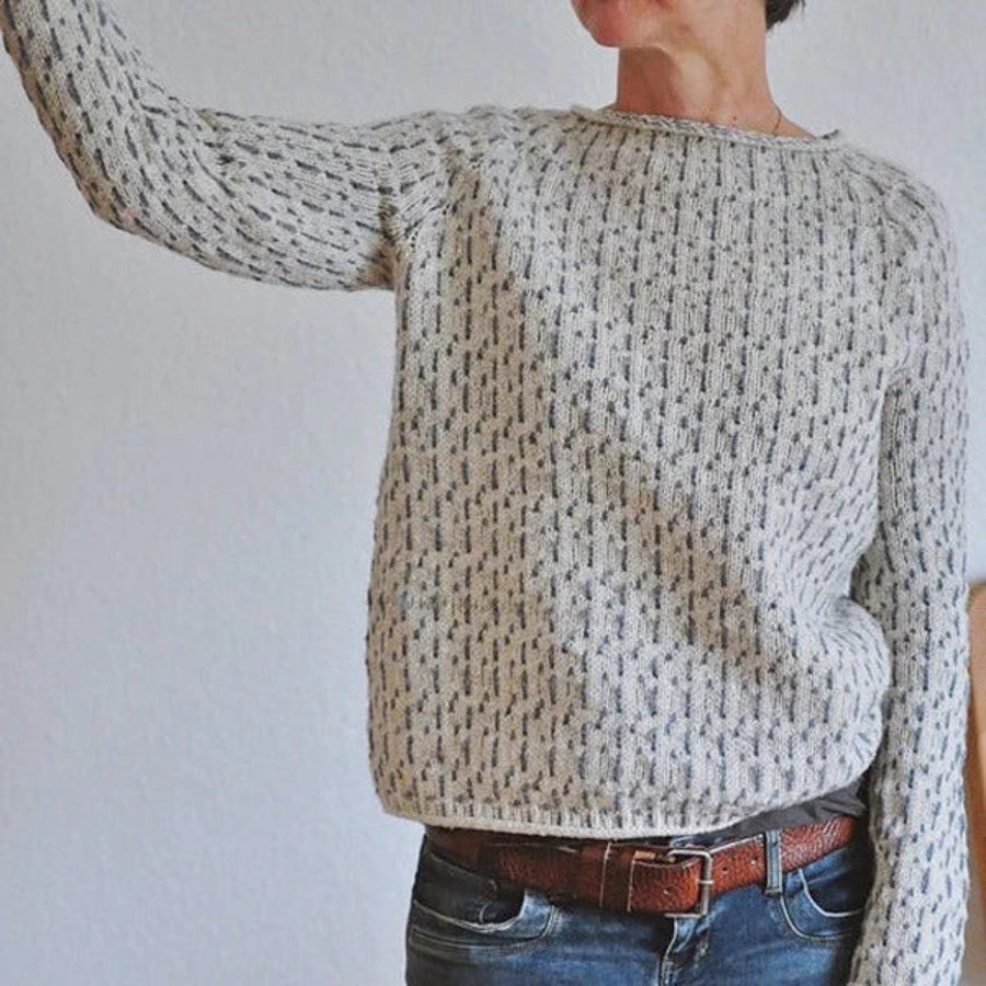 Femme portant un pull en maille clair à col rond, présentant un motif texturé discret et une coupe décontractée, associé à un jean et prise en photo dans un intérieur lumineux.