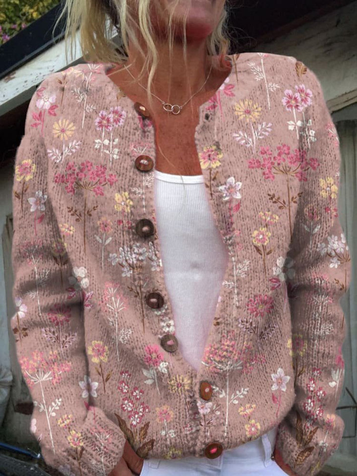 Femme portant un cardigan en maille rose orné de broderies florales multicolores, avec une coupe moderne et des boutons en bois sur le devant, posant en extérieur.