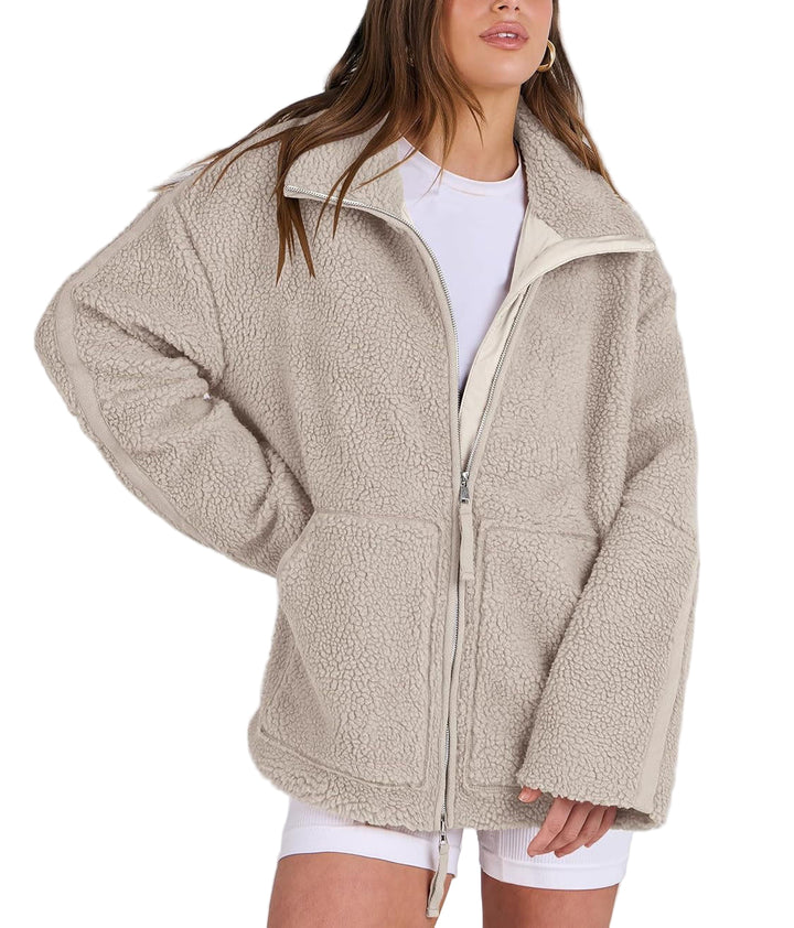 Une femme porte une veste polaire beige, confortable et tendance, avec une coupe ample et un zip central. Le col est légèrement relevé pour plus de chaleur, et la veste est dotée de poches sur le devant pour un look pratique et stylé. Elle est associée à une tenue décontractée, composée d'un t-shirt blanc et de shorts, créant un style à la fois chic et confortable, parfait pour les journées fraîches.