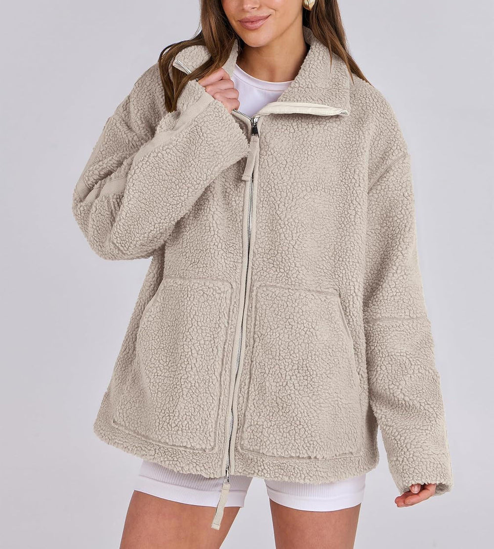 Une femme porte une veste polaire beige à la coupe ample et moderne, avec un zip central et des poches pratiques sur le devant. Elle a un sourire détendu et porte une tenue casual avec un t-shirt blanc et un short, parfait pour un look hivernal confortable et tendance. La texture douce de la veste est bien visible, offrant chaleur et style pour les journées fraîches.