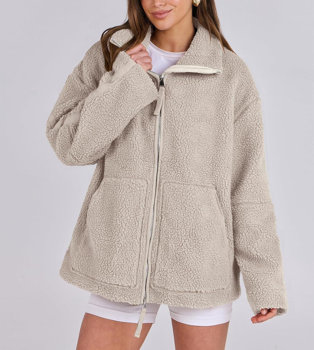 Une femme porte une veste polaire beige à la coupe ample et moderne, avec un zip central et des poches pratiques sur le devant. Elle a un sourire détendu et porte une tenue casual avec un t-shirt blanc et un short, parfait pour un look hivernal confortable et tendance. La texture douce de la veste est bien visible, offrant chaleur et style pour les journées fraîches.