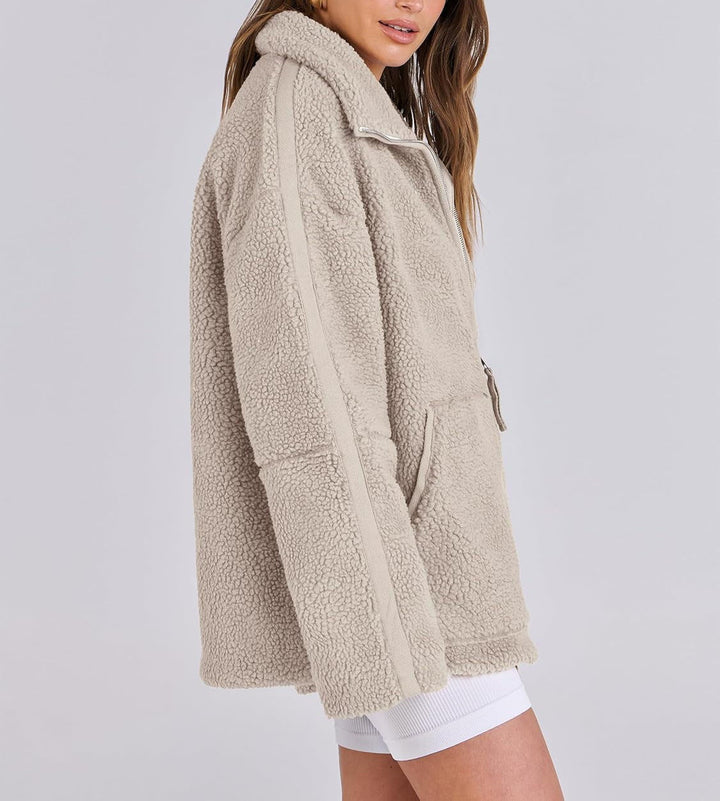 Une femme porte une veste polaire beige, à la coupe ample et moderne. Le col est relevé pour plus de chaleur, et la veste dispose de poches sur le devant, avec une fermeture à zip. Elle est vue de profil, montrant les détails des lignes latérales contrastées sur les manches, apportant une touche de style supplémentaire à ce look tendance et confortable, parfait pour l’hiver.