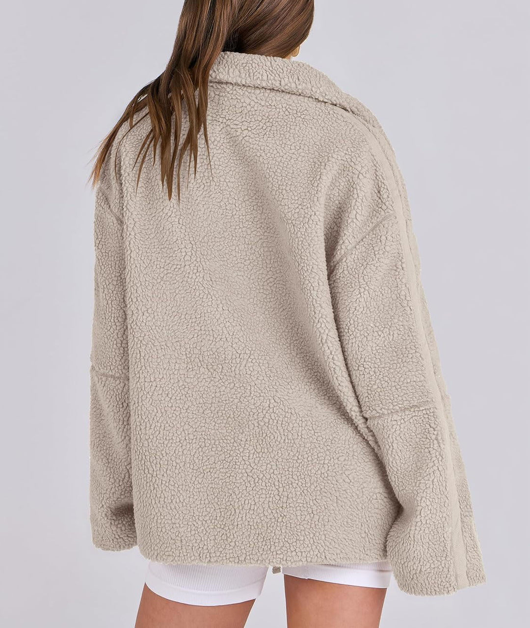 Une femme porte une veste polaire beige vue de dos, montrant sa coupe ample et ses détails de couture contrastés le long des manches. Le col est légèrement relevé pour plus de confort, et la veste présente une texture douce et épaisse, idéale pour les journées fraîches. La silhouette décontractée et stylée de la veste offre une touche moderne pour un look hivernal à la fois tendance et pratique.