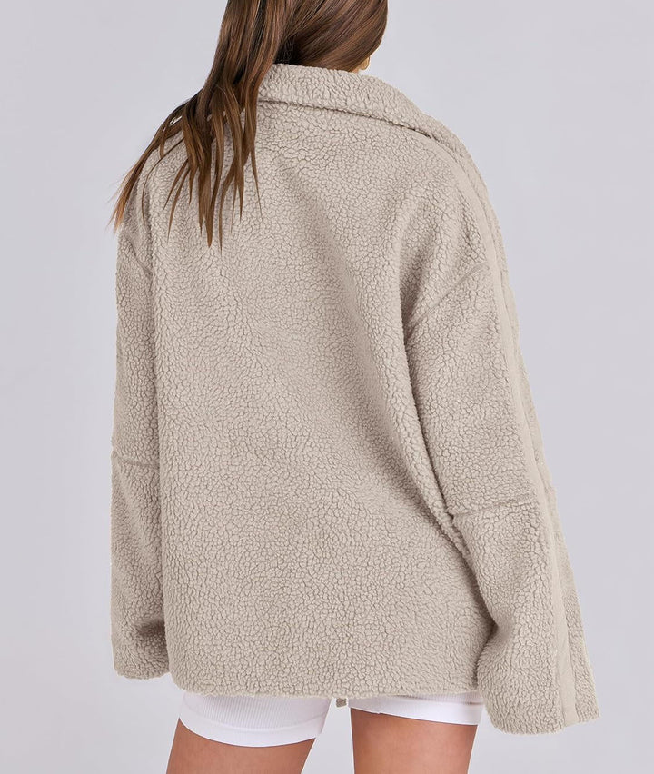 Une femme porte une veste polaire beige vue de dos, montrant sa coupe ample et ses détails de couture contrastés le long des manches. Le col est légèrement relevé pour plus de confort, et la veste présente une texture douce et épaisse, idéale pour les journées fraîches. La silhouette décontractée et stylée de la veste offre une touche moderne pour un look hivernal à la fois tendance et pratique.
