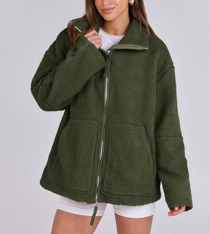 Une femme porte une veste polaire verte, avec une coupe ample et moderne. Le zip central est visible, et les poches sur le devant apportent une touche pratique et stylée. Le col est légèrement relevé pour plus de confort et de chaleur. Elle associe la veste à un t-shirt blanc et un short, créant un look décontracté et tendance, parfait pour les journées fraîches.