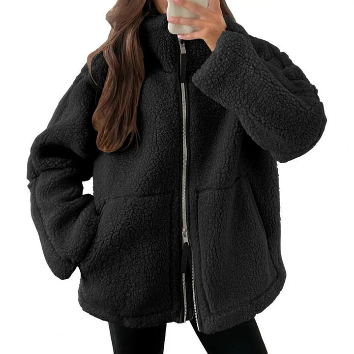 Une femme porte une veste polaire noire avec une coupe ample et moderne. La veste est fermée par un zip central et dispose de poches latérales pour plus de praticité. Le col est légèrement relevé, offrant confort et chaleur. Elle prend un selfie tout en portant la veste, associée à un pantalon noir, créant un look décontracté et tendance, parfait pour les journées froides.