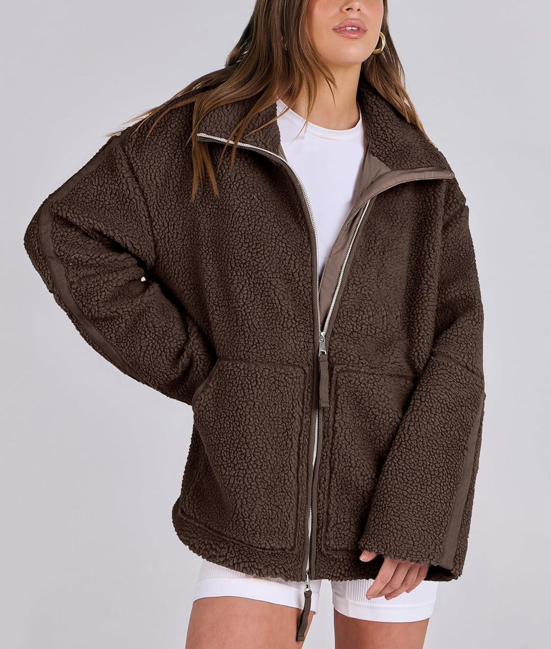 Une femme porte une veste polaire marron avec une coupe ample et moderne. Le col est légèrement relevé pour plus de chaleur et de confort, tandis que le zip central permet de fermer la veste. Les poches sur le devant ajoutent une touche pratique. Elle porte cette veste avec un t-shirt blanc et un short, créant un look décontracté et stylé, parfait pour les journées fraîches.