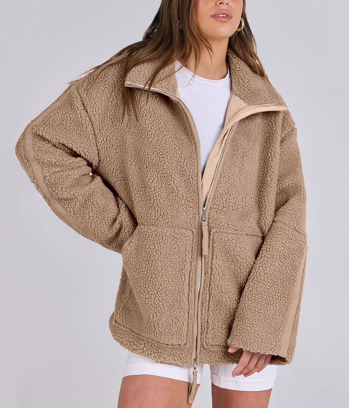 Une femme porte une veste polaire marron clair à la coupe moderne et décontractée. La fermeture éclair centrale est bien visible et le col est légèrement relevé pour plus de confort. La veste possède des poches avant pratiques. Elle l'associe à un t-shirt blanc et un short, créant ainsi un look à la fois décontracté et élégant, idéal pour les journées fraîches. Les détails contrastés des manches et les surpiqûres apportent une touche d'originalité à la tenue.