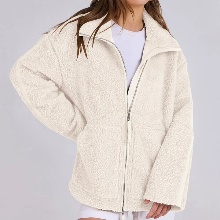 Une femme porte une veste polaire blanche à la coupe ample et moderne, avec un zip central et des poches avant pratiques. Le col est légèrement relevé pour ajouter du confort et de la chaleur. Elle associe la veste à un t-shirt blanc et un short, créant un look décontracté et élégant. La texture douce de la veste en fait un choix parfait pour les journées fraîches.