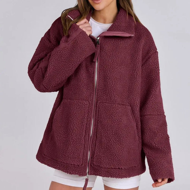 Une femme porte une veste polaire rouge à la coupe décontractée et moderne. La veste est dotée d'une fermeture éclair centrale et de poches avant pratiques. Son col légèrement montant offre un confort et une chaleur appréciables. Elle porte un t-shirt blanc et un short, pour un look à la fois décontracté et élégant. La douceur de la veste contribue à son confort, la rendant idéale pour les journées fraîches.