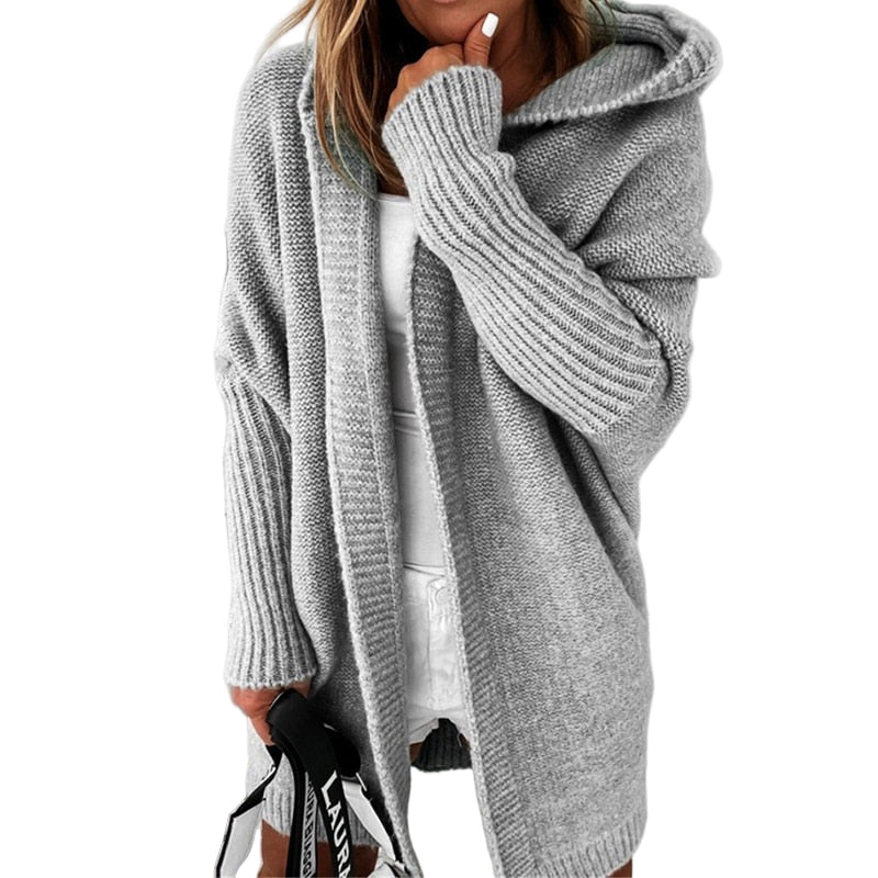 Femme portant un long cardigan gris à maille épaisse avec capuche, superposé à une tenue blanche, tenant un sac à sangle imprimée, dans un décor minimaliste aux tons clairs.