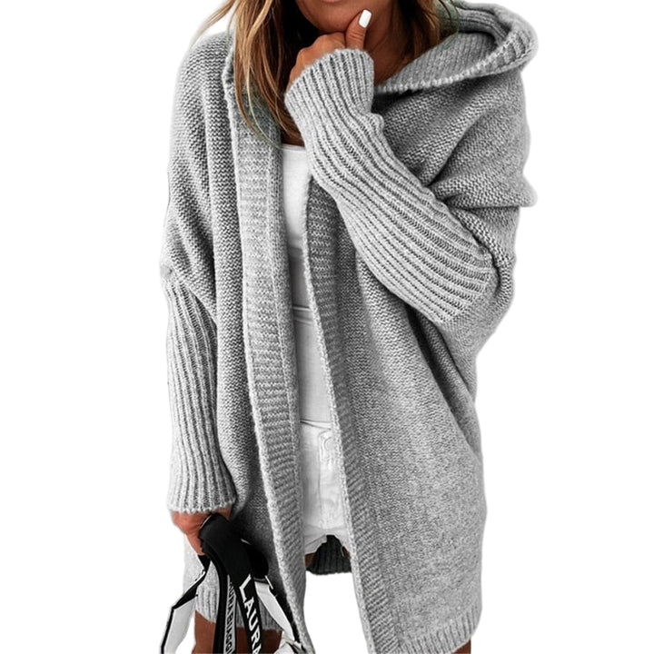 Femme portant un long cardigan gris à maille épaisse avec capuche, superposé à une tenue blanche, tenant un sac à sangle imprimée, dans un décor minimaliste aux tons clairs.