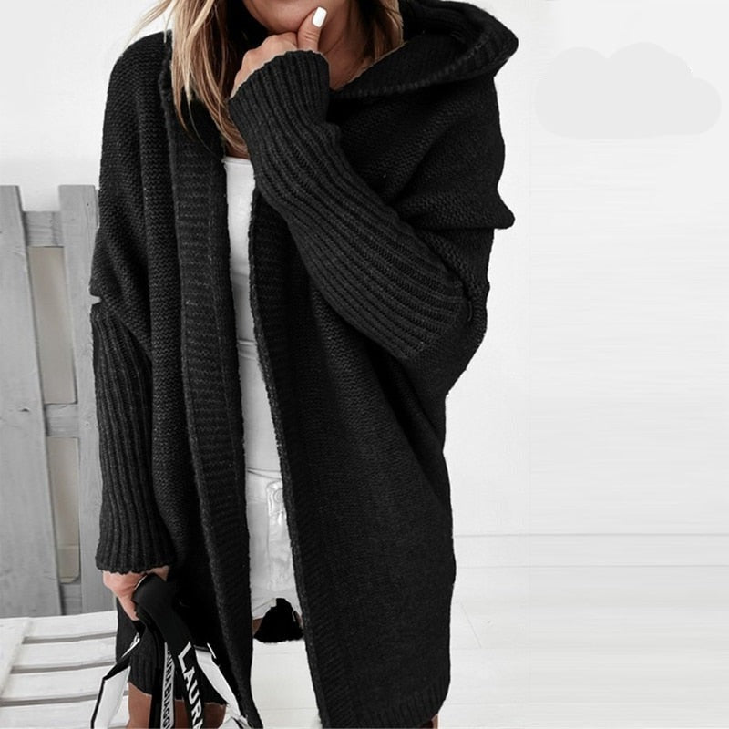 Femme portant un long cardigan noir à maille épaisse avec capuche, superposé à une tenue blanche, tenant un sac à sangle imprimée, dans un décor lumineux et minimaliste.