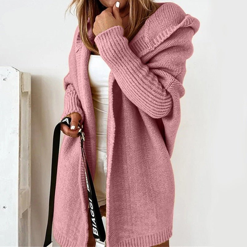 Femme portant un long cardigan rose à maille épaisse avec capuche, superposé à une tenue blanche, tenant un sac à sangle imprimée, dans un intérieur lumineux au style minimaliste.