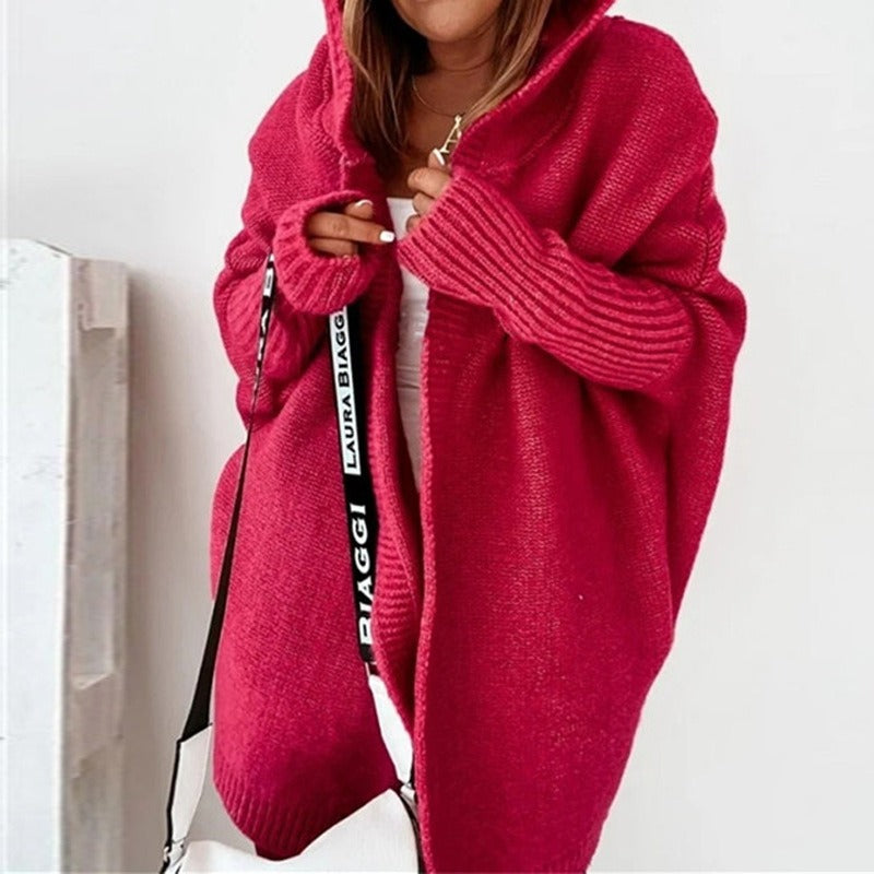 Femme portant un long cardigan rouge en maille épaisse à capuche, superposé à une tenue blanche, tenant un sac à bandoulière à bandoulière imprimée, dans un intérieur lumineux de style minimaliste.