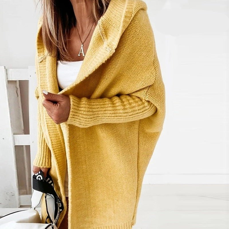 Femme portant un long cardigan jaune à maille épaisse avec capuche, superposé à un haut blanc, tenant un sac à sangle imprimée, dans un intérieur lumineux au style minimaliste.