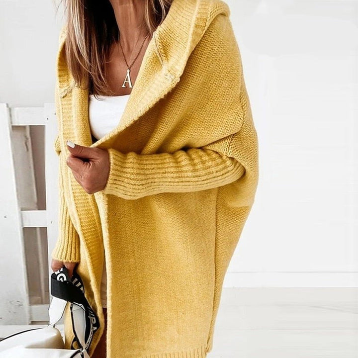Femme portant un long cardigan jaune à maille épaisse avec capuche, superposé à un haut blanc, tenant un sac à sangle imprimée, dans un intérieur lumineux au style minimaliste.