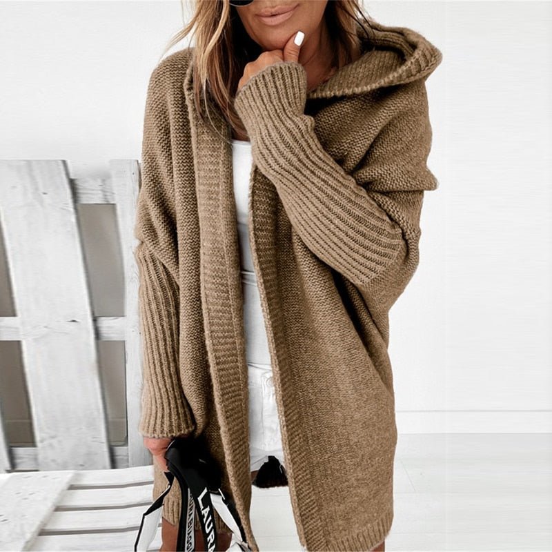 Femme portant un long cardigan marron à maille épaisse avec capuche, superposé à une tenue blanche, tenant un sac à sangle imprimée, dans un intérieur lumineux au style minimaliste.