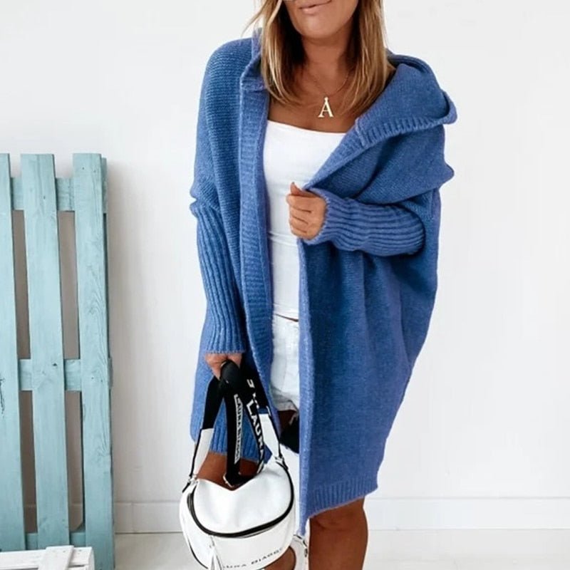 Femme portant un long cardigan bleu à maille épaisse avec capuche, superposé à une tenue blanche, tenant un sac blanc à sangle imprimée, dans un intérieur lumineux au style minimaliste.