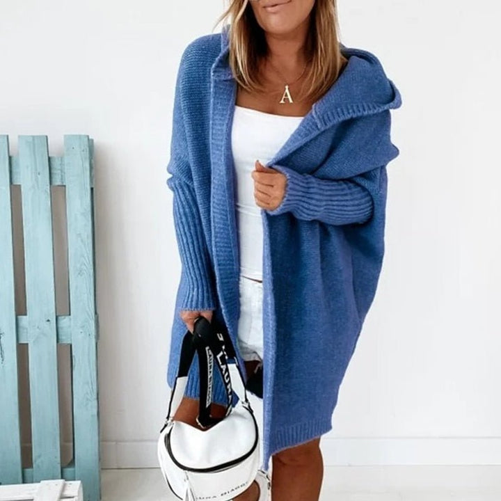 Femme portant un long cardigan bleu à maille épaisse avec capuche, superposé à une tenue blanche, tenant un sac blanc à sangle imprimée, dans un intérieur lumineux au style minimaliste.
