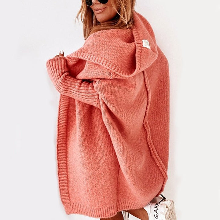 Femme de dos portant un long cardigan corail à maille épaisse avec capuche, associé à une tenue claire et un sac blanc, posant dans un intérieur lumineux au style minimaliste.