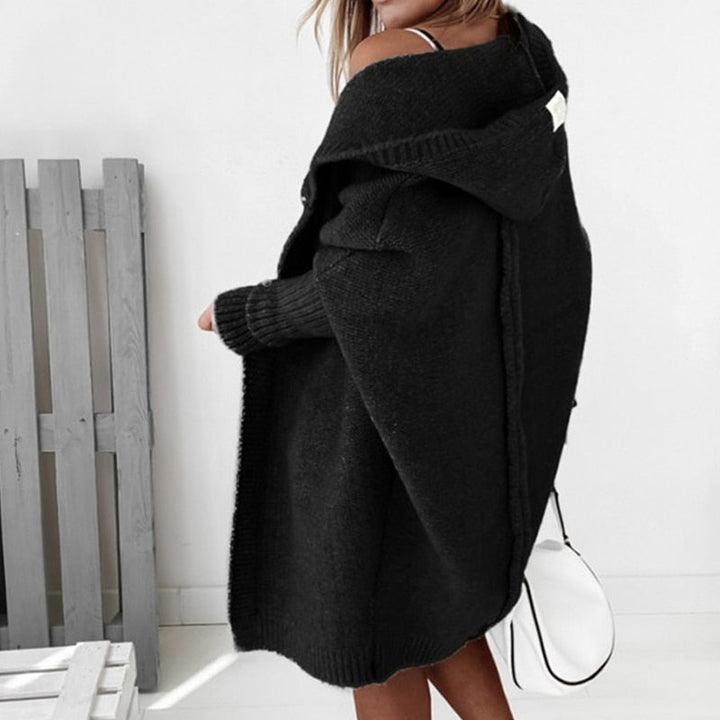 Femme vue de dos portant un long cardigan noir à maille épaisse avec capuche, superposé à une tenue claire, tenant un sac blanc, dans un intérieur lumineux au style minimaliste.